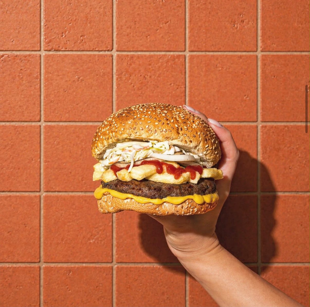 Burger sésame tenu à la main devant un carrelage terracotta